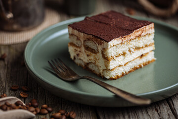 A piece of Tiramisu cake, traditional Italian dessert, served in moody, rustic scene. Wooden background and green plate. 
