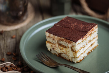 A piece of Tiramisu cake, traditional Italian dessert, served in moody, rustic scene. Wooden background and green plate. 