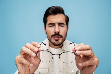Close-up portrait of a brunette man looking through glasses that he holds in his hands, eye problems, glasses for vision farsightedness and nearsightedness, on a blue background, copy space 