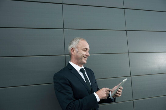Mature businessman standing in front of wall, looking at tablet computer