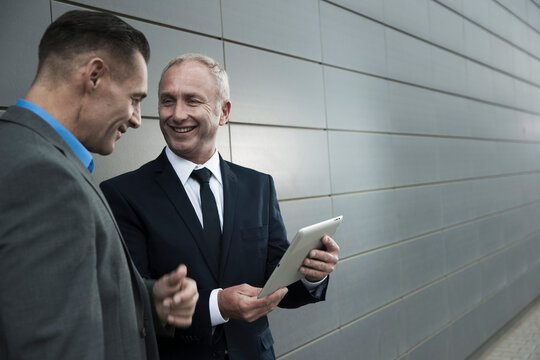 Mature businessmen standing in front of wall, talking and looking at tablet computer