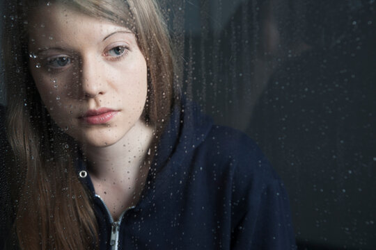 Portrait of young woman behind window, wet with raindrops, wearing hoodie