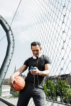 Mature man standing on outdoor basketball court holding basketball and looking at MP3 player, Germany