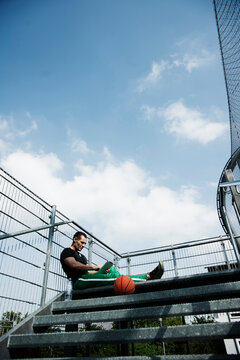 Mature man sitting at top of stairs on outdoor basketball court looking at tablet computer, Germany