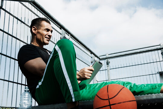 Mature man sitting at top of stairs on outdoor basketball court looking at tablet computer, Germany