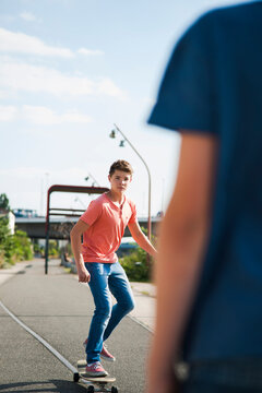 Teenage boy skateboarding on road with back of another boy in foreground, Germany