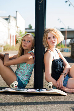 Portrait of teenage girls outdoors with skateboard, looking at camera, sitting on street, Germany