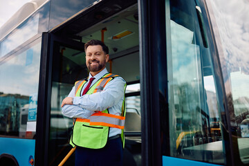 Happy bus driver with arms crossed standing at vehicle entrance door and looking at the camera.