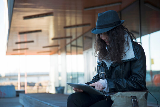 Teenage girl sitting outdoors at train station, wearing fedora and looking at tablet computer, Mannheim, Germany