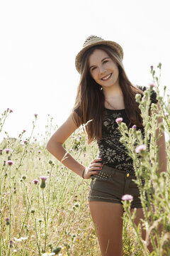 Portrait of teenage girl wearing shorts and straw hat standing in field, smiling and looking at camera, Germany