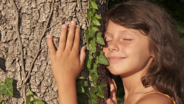 Catch sun against tree trunk. A little girl with close eyes stay by the oak tree trunk during sunny day. A concept of ecological global problems.