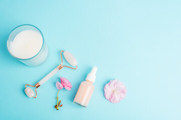 Serum bottle, face roller, gua sha stone, aroma candle and pink sakura blossom on blue background. Skin care, beauty treatment concept. Top view, flat lay