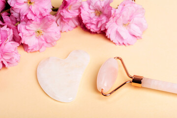 Face roller, gua sha stone and pink cherry blossom on beige background. Skin care, beauty treatment concept. Top view