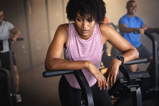 Tired biracial young woman with afro hair exercising on bike in gym