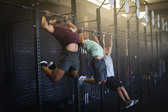 Full length of dedicated biracial young men practicing pull ups on bars in gym