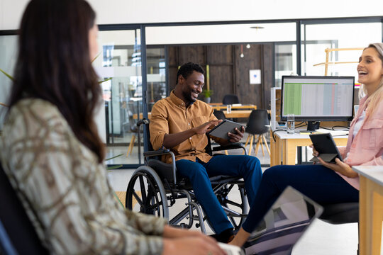 Diverse man sitting on wheelchair and using tablet while discussing with female coworkers in office