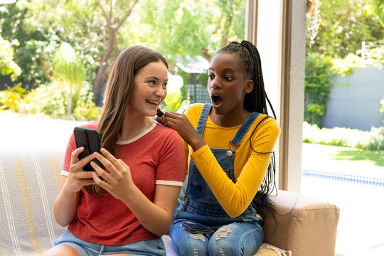 Happy diverse teenager girls friends sitting on sofa, braiding hair and using smartphone