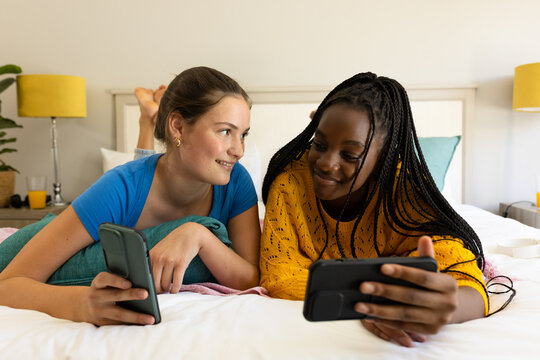 Happy diverse teenager girls friends lying on bed, using smartphones and talking