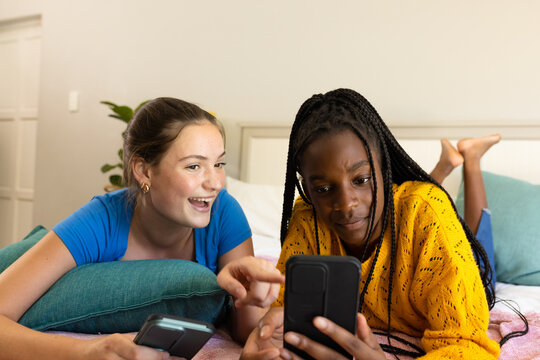 Happy diverse teenager girls friends lying on bed, using smartphones and talking
