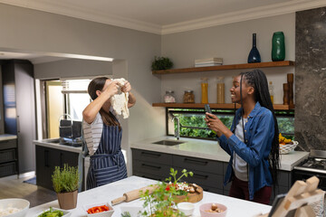Happy diverse teenager girls friends making pizza and using smartphone in kitchen