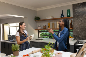 Happy diverse teenager girls friends making pizza and using smartphone in kitchen