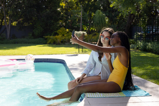 Happy diverse teenager girls friends sitting by swimming pool and taking selfie