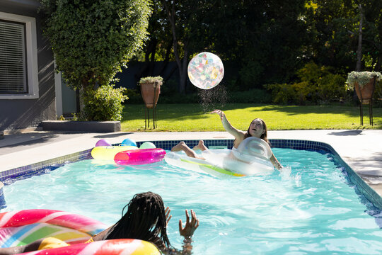 Happy diverse teenager girls friends lying on pool floats and playing in swimming pool in garden