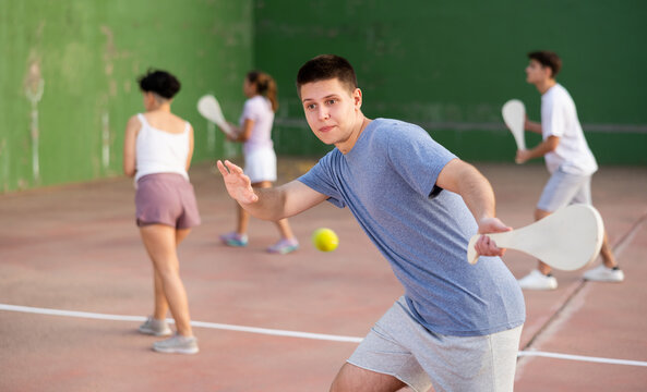 Focused young man playing friendly pelota goma match on outdoor summer court with fronton wall, swinging wooden paleta to hit small rubber ball