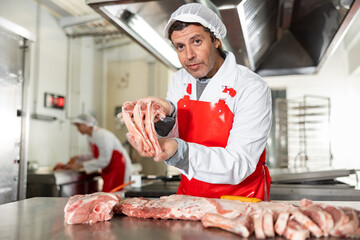 Confident adult butcher in white uniform and red apron standing at meat cutting table, offering fresh raw lamb ribs for sale