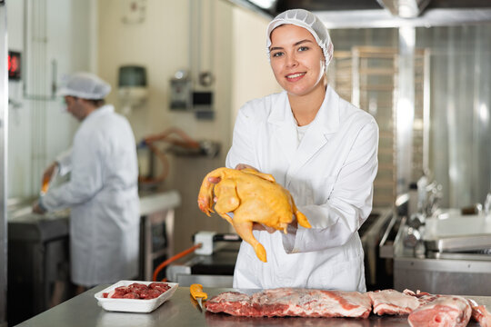 Positive successful young female worker of butchery standing at cutting table in meat processing workshop, showing fresh raw dressed chicken..