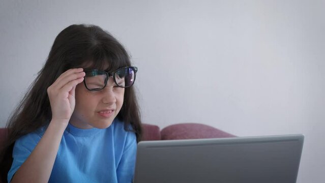Child with glasses squinting eyes by laptop. A stressed little girl wear glasses for better vision by her laptop. A concept of eyes trouble because of computer working at home.