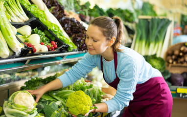 Positive middle-aged saleswoman putting fresh cauliflowers on food stall in grocery store
