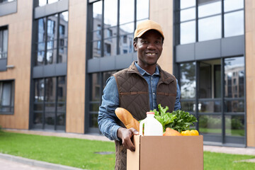 Waist up portrait of black young man carrying box with fresh vegetables while delivering groceries...