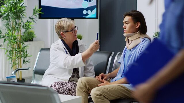 Senior medic doing concussion exam to check asian patient vision and brain damage with light. Person with neck collar doing consultation with elderly physician in hospital waiting area