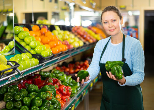 Portrait Of Busy European Female Seller In Apron Arranging Bell Peppers On Shelves In Grocery Supermarket