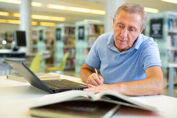 Fototapeta premium Concentrated mature man spending time in library, reading books and making laptop. Self-education concept