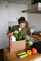 Vertical portrait of young woman unpacking fresh grocery delivery in kitchen - obrazy, fototapety, plakaty