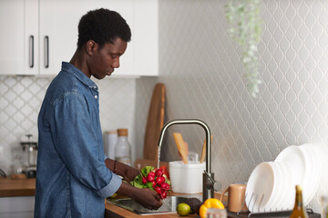 Side view portrait of black young man washing fresh vegetables in kitchen, copy space