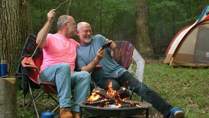Two gay men with pride flag and tent in front of campfire talking to smartphone on video chat or taking selfie and kiss.