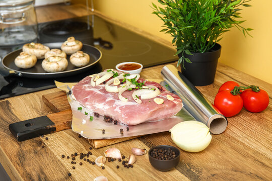 Aluminium foil roll with raw piece of meat, spices and vegetables on wooden table in kitchen