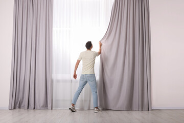 Man closing window curtains at home, back view