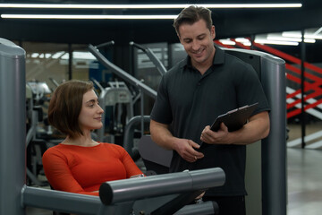 Fototapeta na wymiar Young Caucasian woman doing workout in the gym for her strength with the advisory from her trainer.She is wearing red exercise top outfit and and hands on the workout machine.