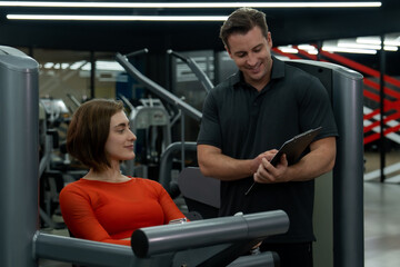 Young Caucasian woman doing workout in the gym for her strength with the advisory from her trainer.She is wearing red exercise top outfit and and hands on the workout machine.