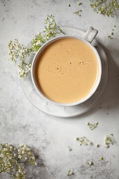 A latte style coffee drink in a mug with a frothy top. The mug is surrounder by white flowers.