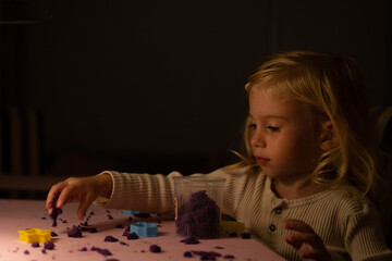 Little two or three year old girl playing with coloured kinetic sand at home in evening in darkness with lamp at table.Kid sensory motor skills development.