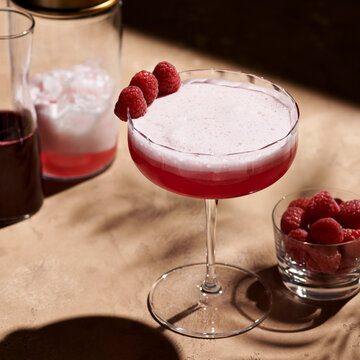 Clover club cocktail in a tall coupe glass garnished with three raspberries next to a shaker in hard light.