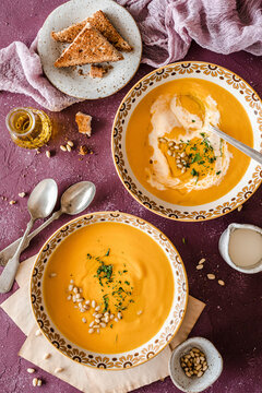 Two bowls of pumpkin soup served with brown toasted bread and garnished with cream, pine nuts and herbs on a purple background