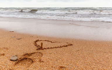 The seas are full on the sandy shore on a cloudy day. A heart is painted on the sand, and the waves wash away the drawing. Rest on the sea. There is an empty space for text