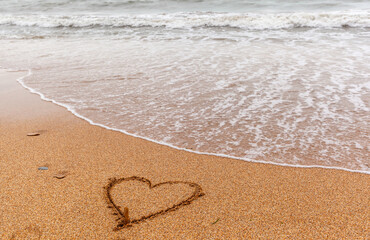 The seas are full on the sandy shore on a cloudy day. A heart is painted on the sand, and the waves wash away the drawing. Rest on the sea. There is an empty space for text