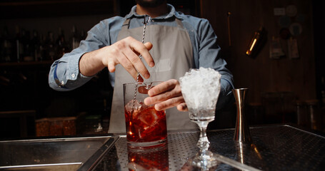Professional bartender masterly creating a cocktail in modern bar. Experienced barman mixing red...